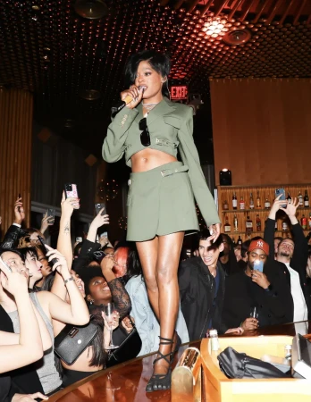 Singer Keke Palmer performs on a bar surrounded by a crowd holding up phones, wearing a green crop top and skirt.