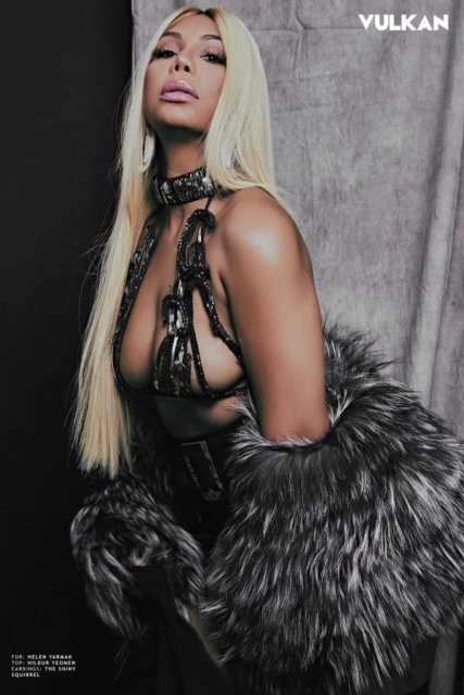 Fashion editorial shot of a woman with platinum blonde hair wearing a metallic-style top and fur wrap against a grey backdrop.
