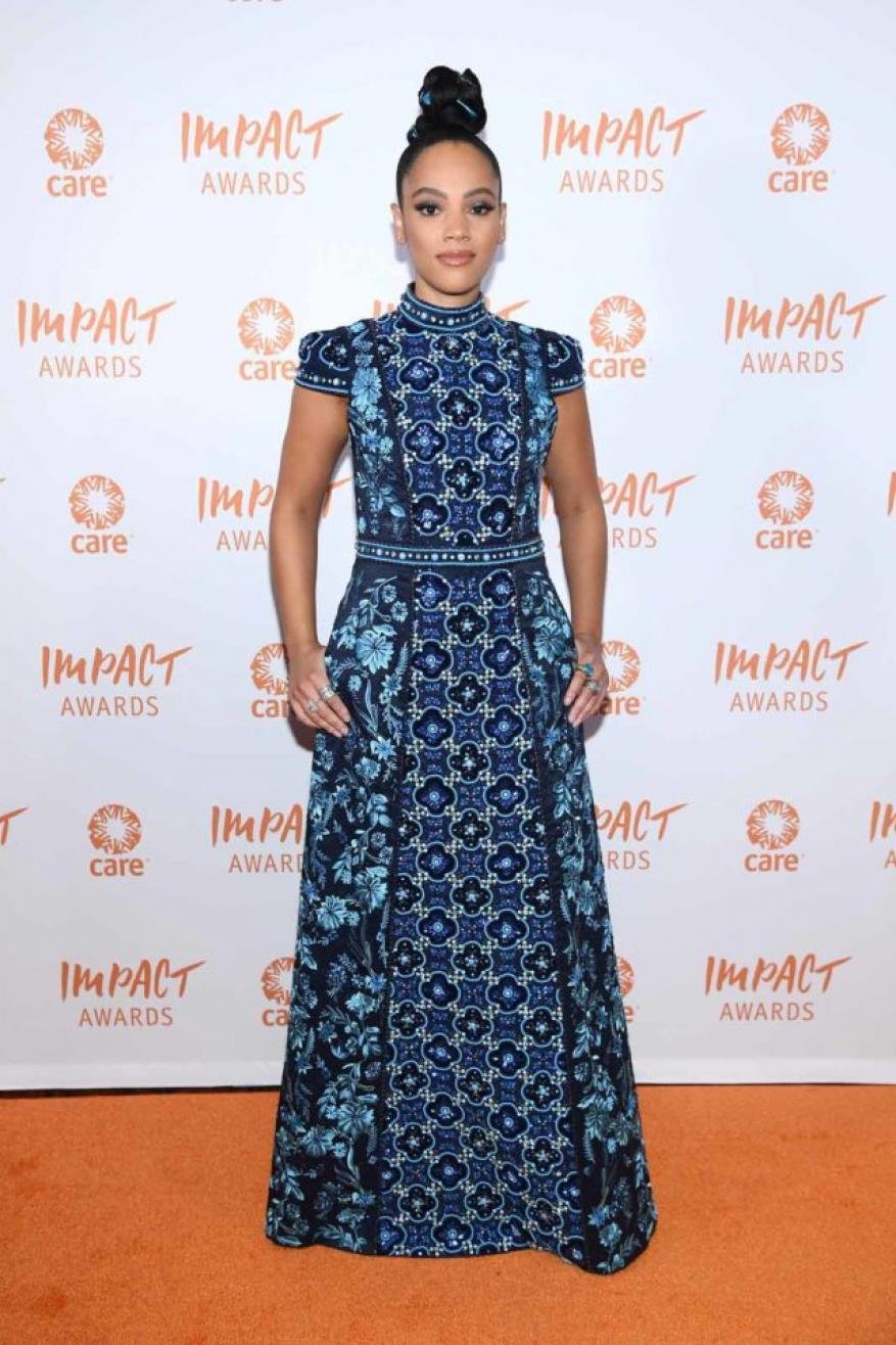 Woman in a patterned blue dress standing on an orange carpet in front of a white backdrop with the Impact Awards logo displayed.