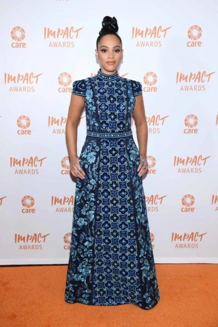 Woman in a patterned blue dress standing on an orange carpet in front of a white backdrop with the Impact Awards logo displayed.