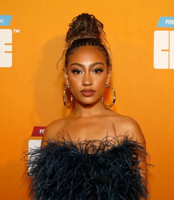Portrait of a stylish young woman with updo, hoop earrings, and a black feather dress against an orange background.