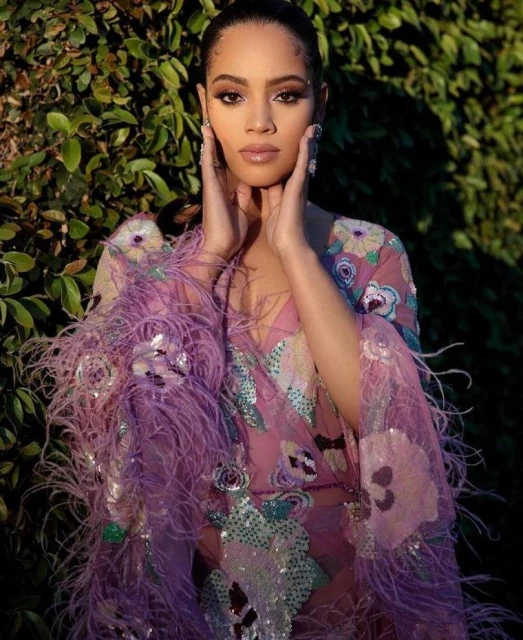 Portrait of a young adult woman with fair skin, wearing a lavender feather-trimmed dress, posing outdoors among green leafy plants.