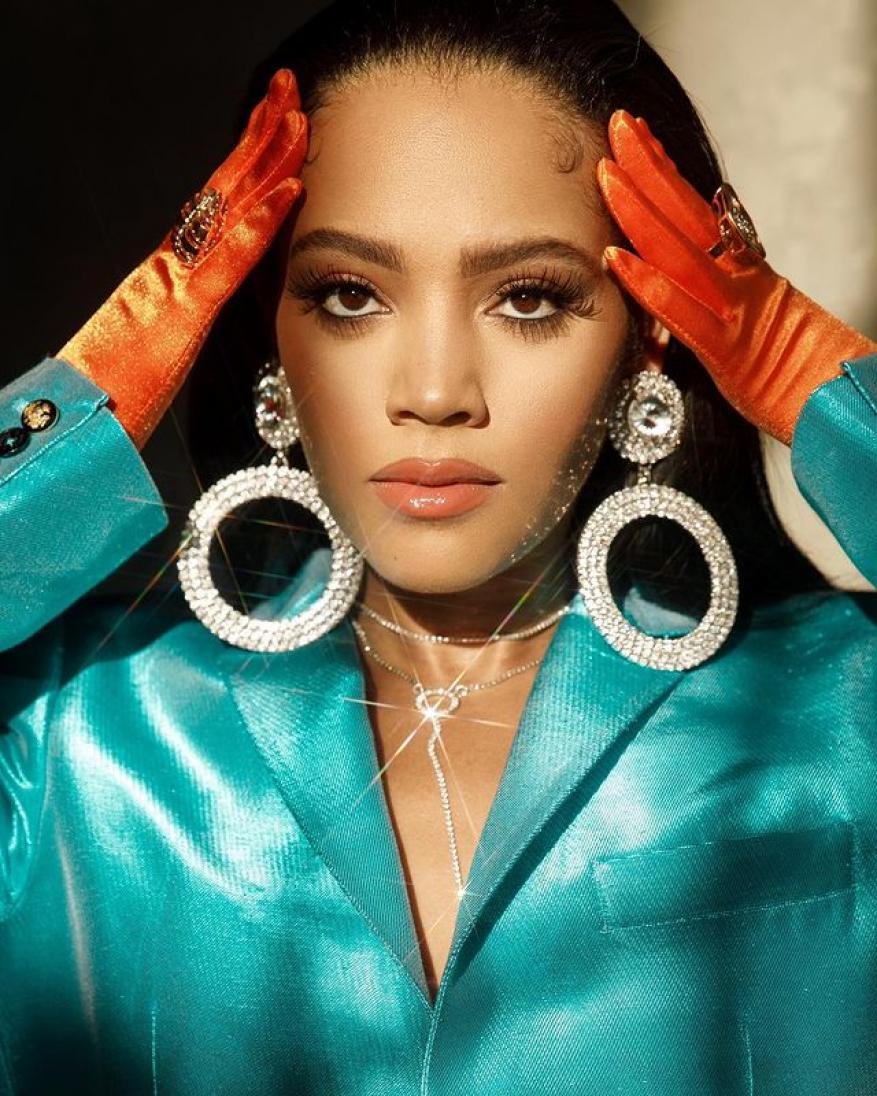 Portrait of a woman with large rhinestone earrings wearing a blue jacket and orange gloves posing indoors with soft, diffused lighting.