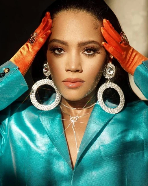 Portrait of a woman with large rhinestone earrings wearing a blue jacket and orange gloves posing indoors with soft, diffused lighting.