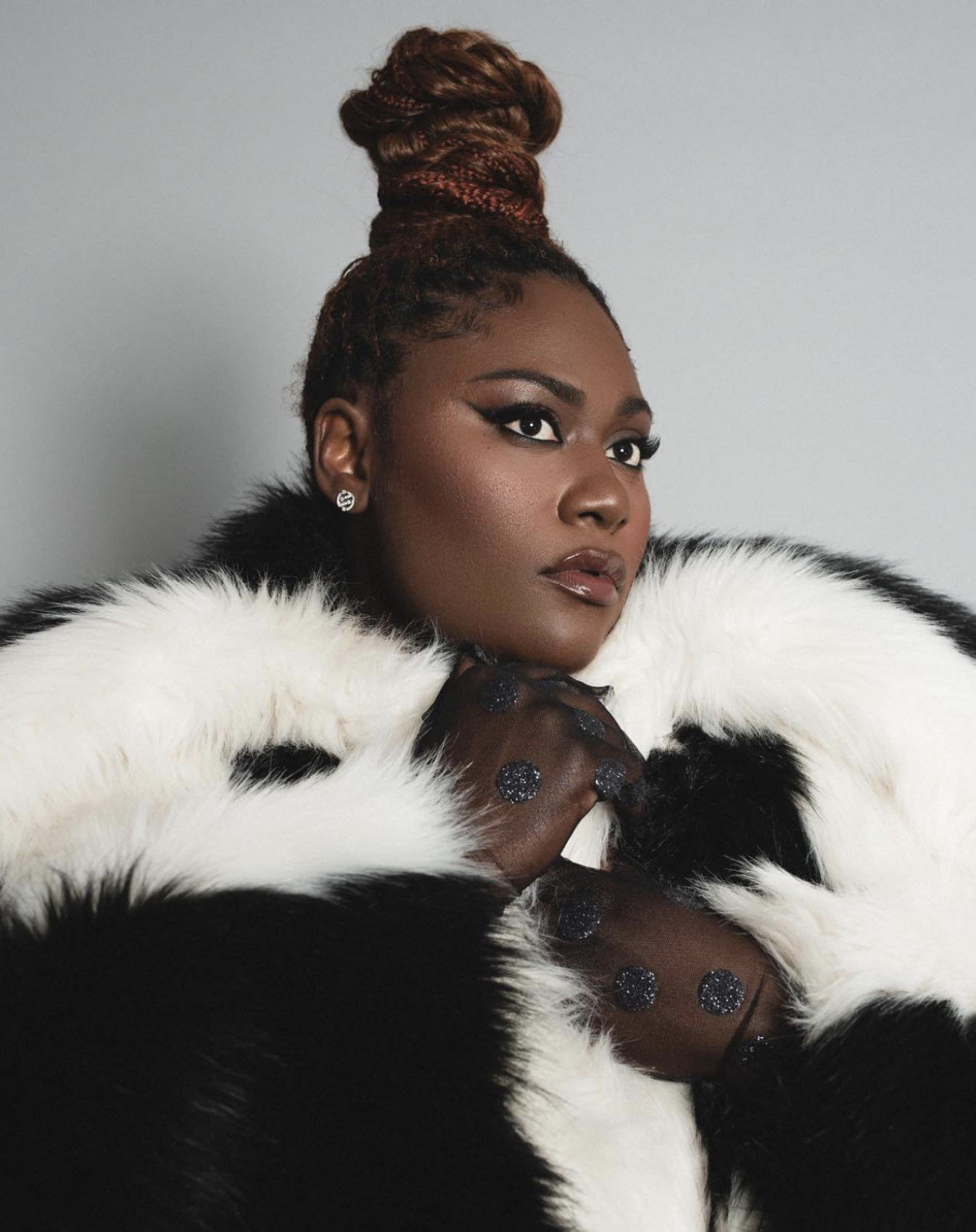 Headshot of a Black female in a black-and-white faux fur coat against a gray background with smokey eye makeup and a high braided bun.