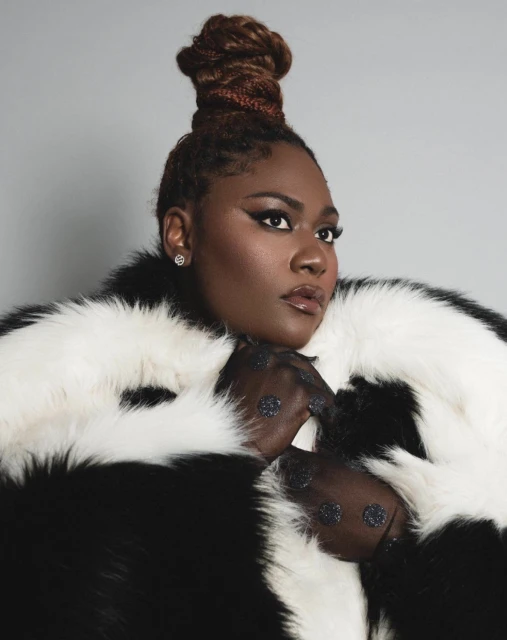 Headshot of a Black female in a black-and-white faux fur coat against a gray background with smokey eye makeup and a high braided bun.