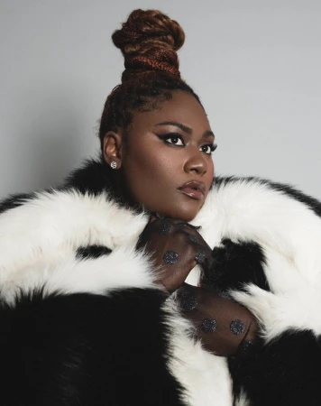 Headshot of a Black female in a black-and-white faux fur coat against a gray background with smokey eye makeup and a high braided bun.