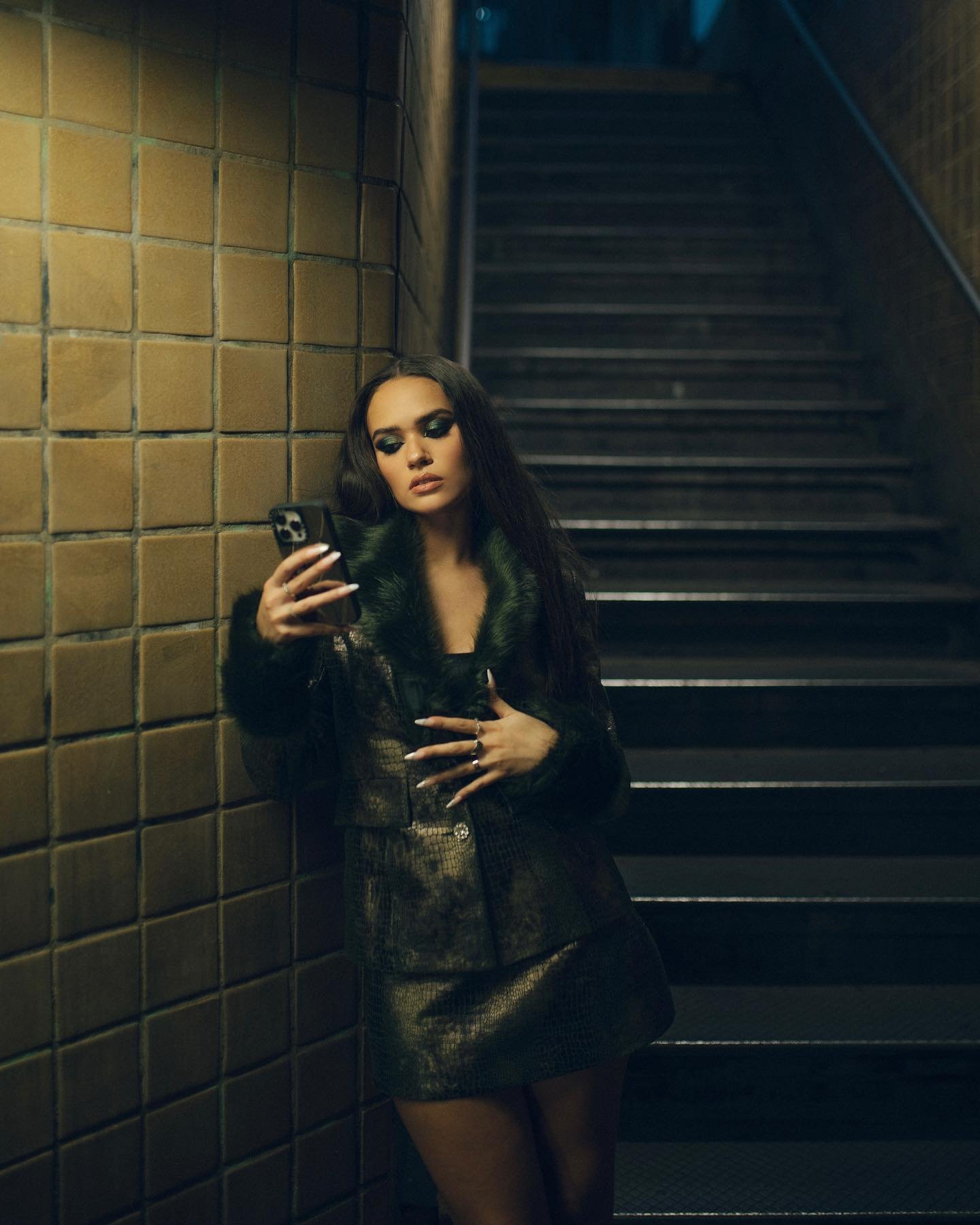 Young woman in a fur-collared coat poses in a stairwell, illuminated by ambient light, holding a phone and looking at the screen.