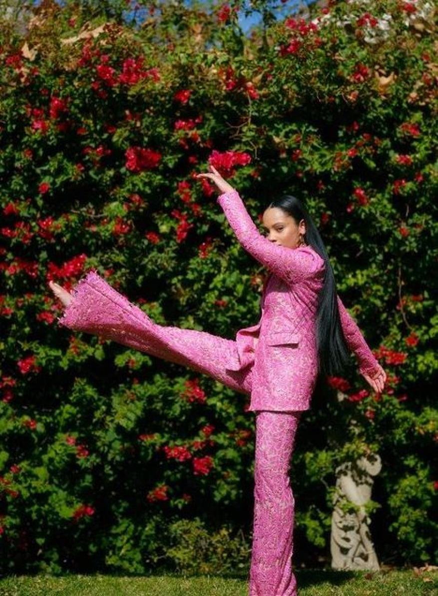 Woman in pink suit poses dynamically in garden with red flowers. Full shot shows dancer on green grass lawn in floral setting.