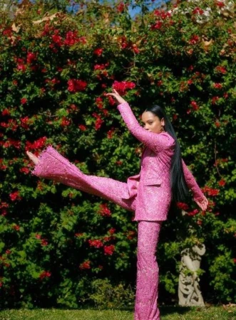 Woman in pink suit poses dynamically in garden with red flowers. Full shot shows dancer on green grass lawn in floral setting.