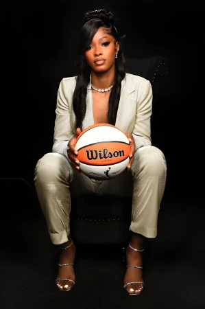Portrait of a woman in a suit holding a basketball in a studio setting. She has dark hair and wears jewelry, posing against a black backdrop.