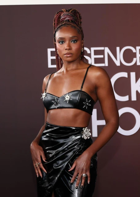 A young adult Black woman poses in a black two-piece outfit with braided hair, standing against a gradient brown backdrop at a red-carpet event.