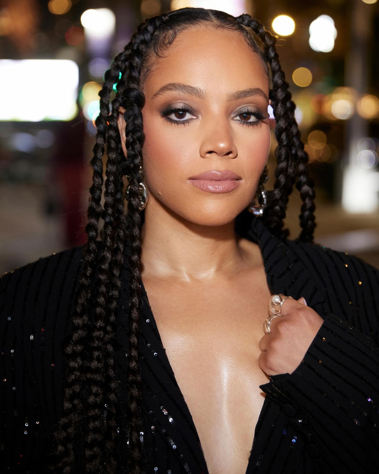 Close-up portrait of a young Black woman with braided hair, wearing a black sequined blazer, shot at eye-level in an outdoor setting at night.