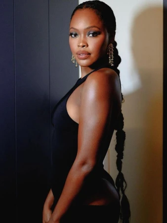 Black woman with long braided hair wearing a black sleeveless top and dangling earrings against a neutral split background.