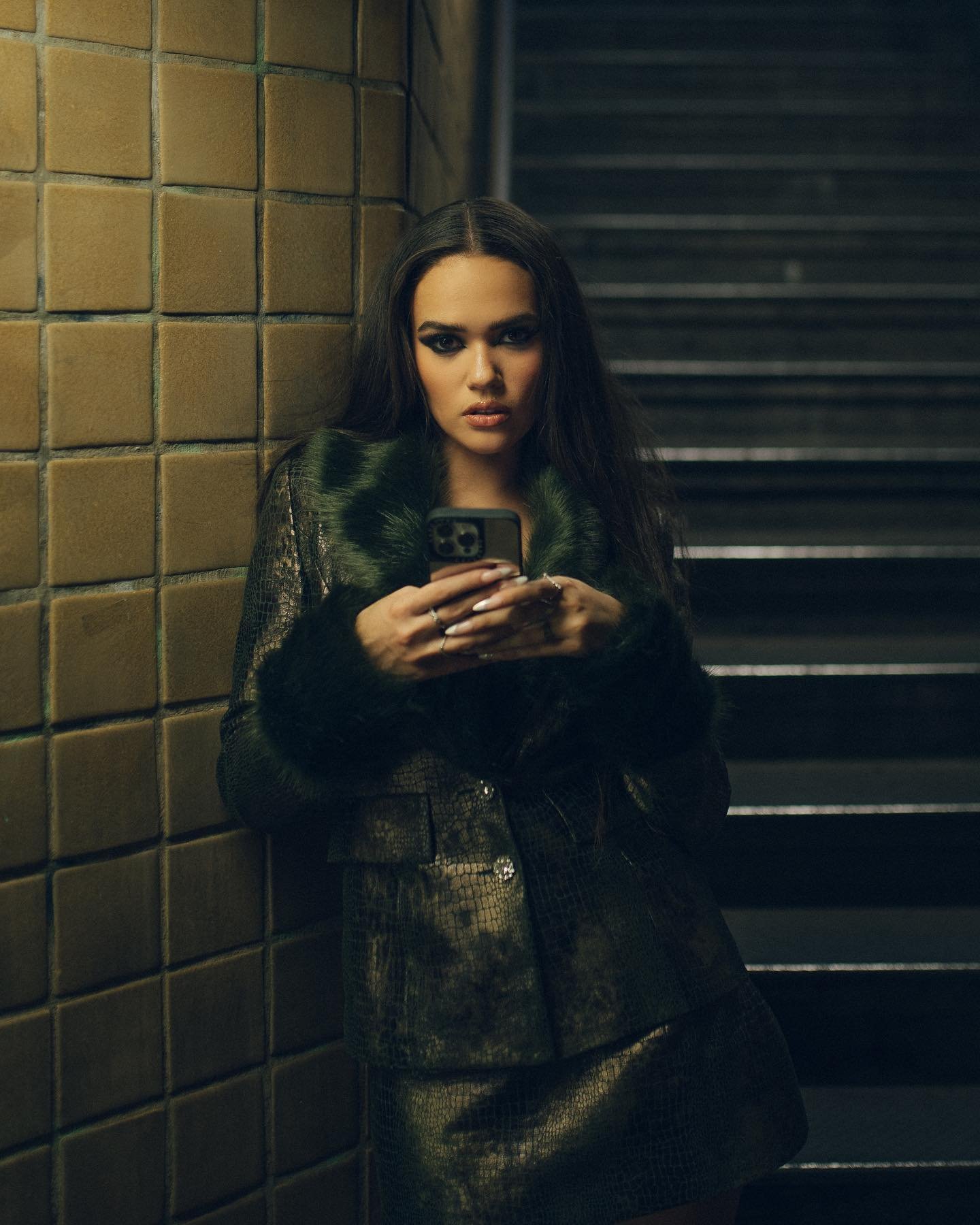 Brunette woman in a green and gold skirt suit leans against a tiled wall, looking at her cell phone on a dark stairwell.