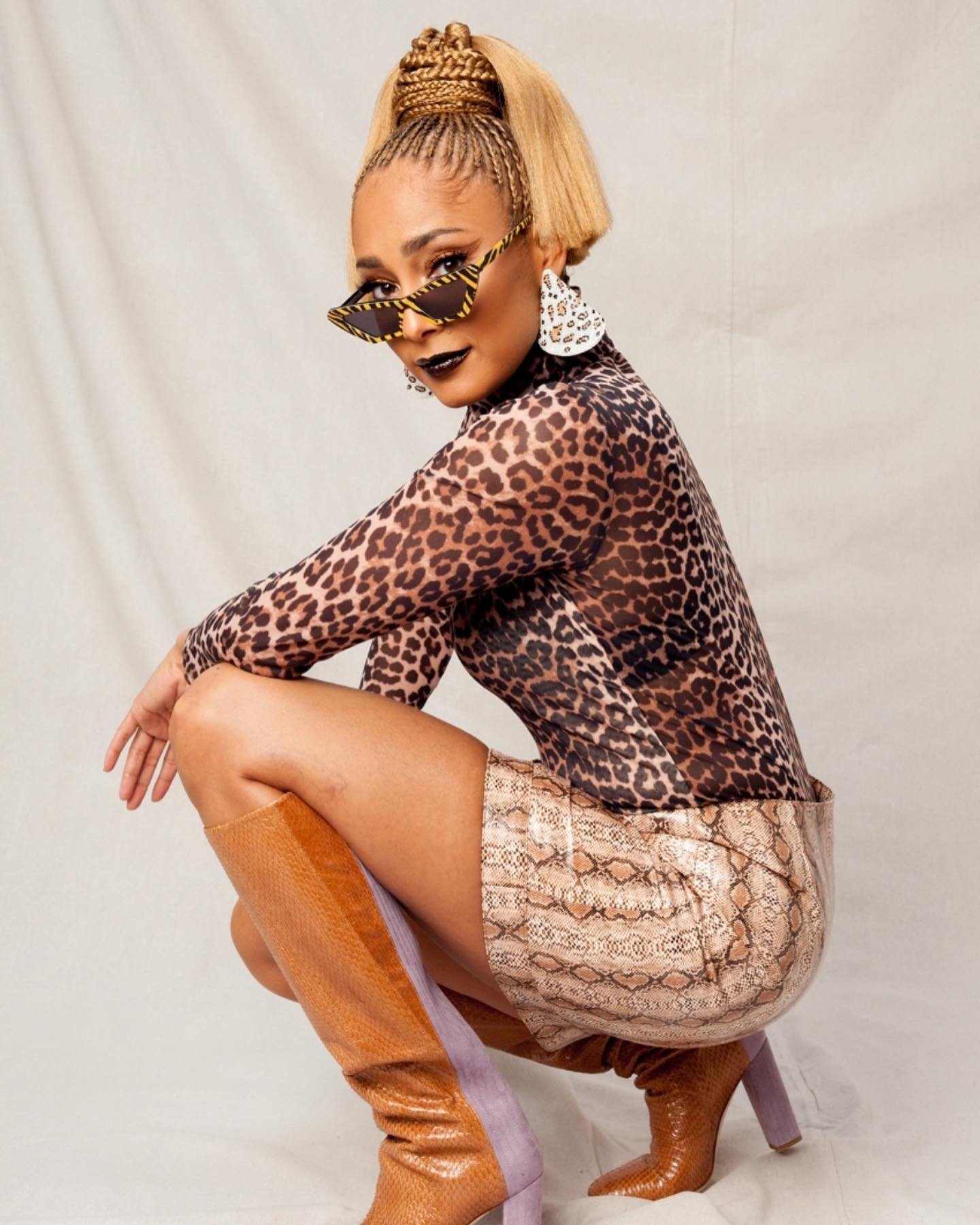 A woman with unique style poses in a studio. She wears a leopard-print top, snakeskin skirt, and brown snakeskin-print knee-high boots.