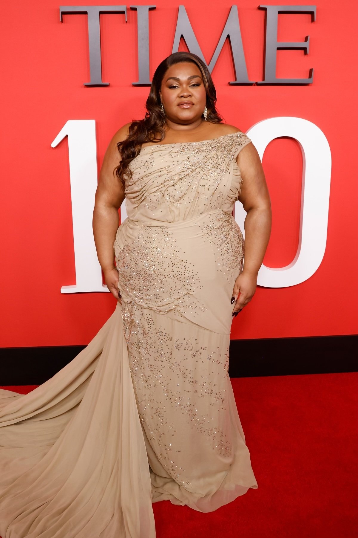 Full-length portrait of a Black female celebrity posing on a red carpet in an elegant, champagne-colored, beaded, one-shoulder gown with a train.
