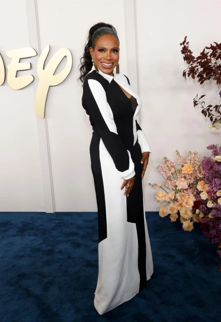 Elegant woman in a black and white gown stands on a blue carpet at a Disney event with floral arrangements in the background.