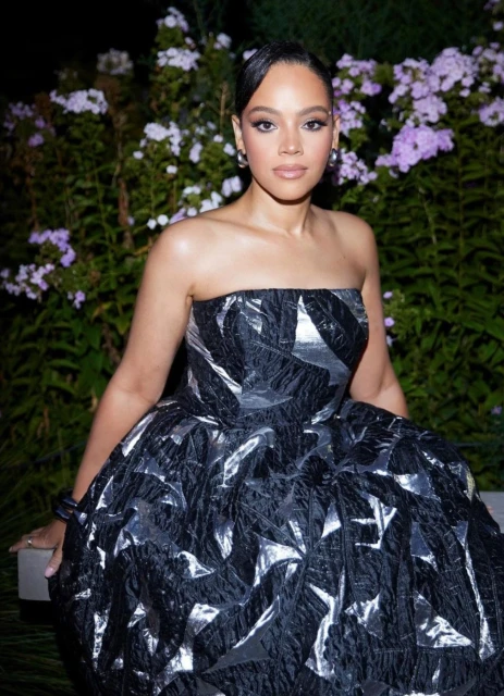 A woman in a strapless black dress poses in front of lush greenery and lavender flowers, captured in a medium shot.