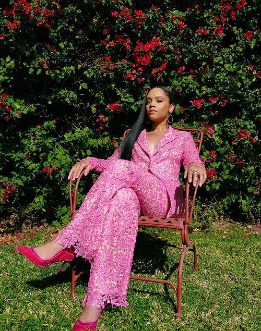 Woman in pink lace suit sitting on a metal chair in a yard with red flowers and green foliage, medium shot, eye-level angle.