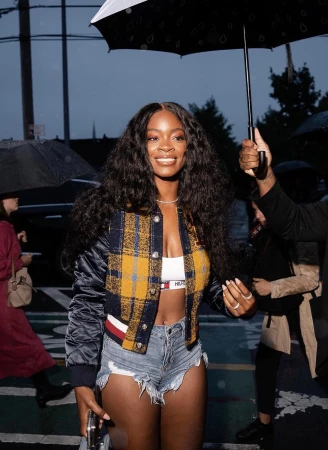 A smiling black woman with long dark wavy hair wearing a yellow plaid jacket, white crop top, and denim shorts under an open black umbrella.