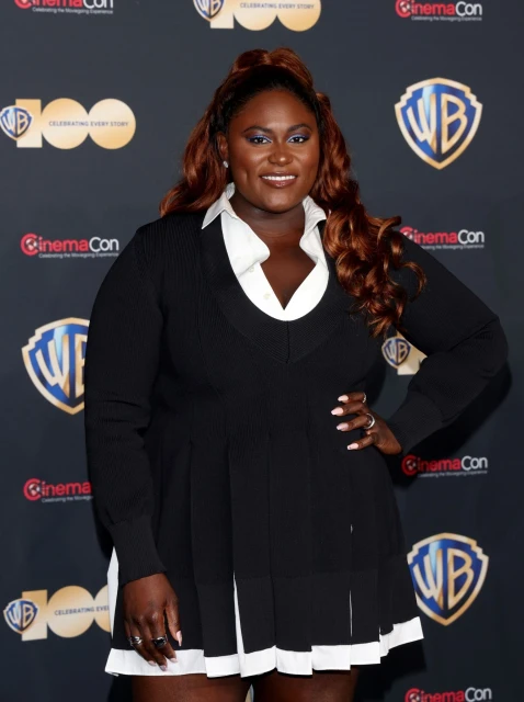 Headshot of a smiling woman with auburn hair wearing a black and white dress, posing in front of a branded CinemaCon backdrop.