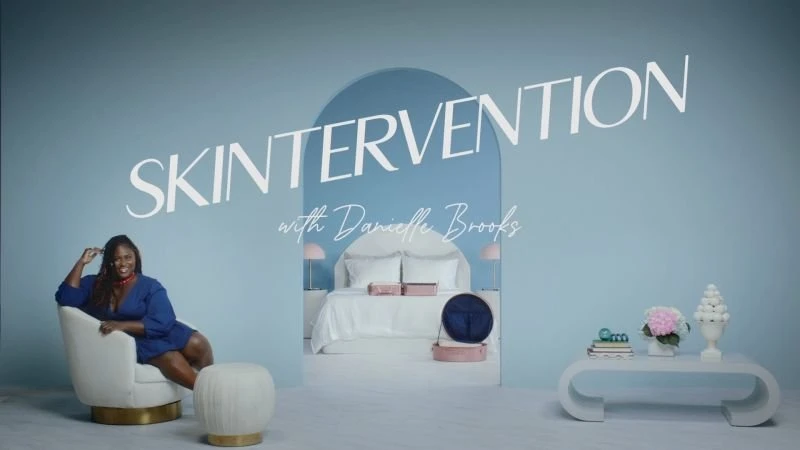 Adult woman in a blue dress is seated in a white chair, in front of a blue background, next to a set with bed and decorations.