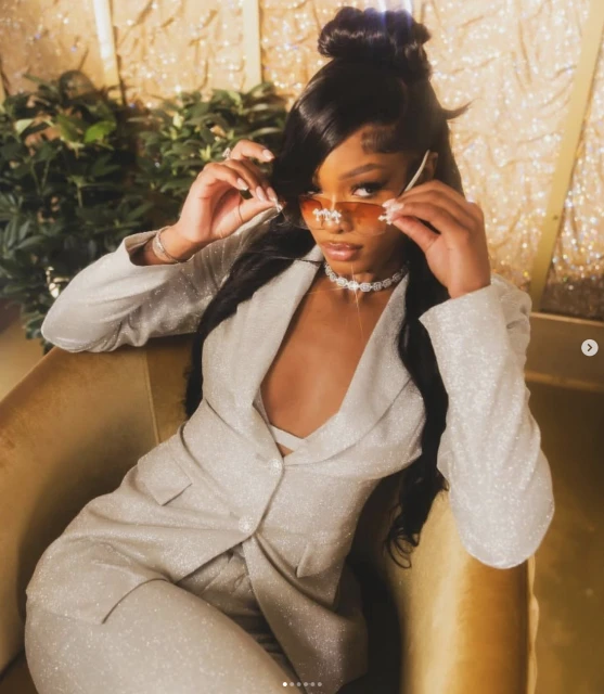 Elegant woman in a sparkling silver suit poses indoors wearing stylish sunglasses and jewelry against a shimmering gold backdrop.