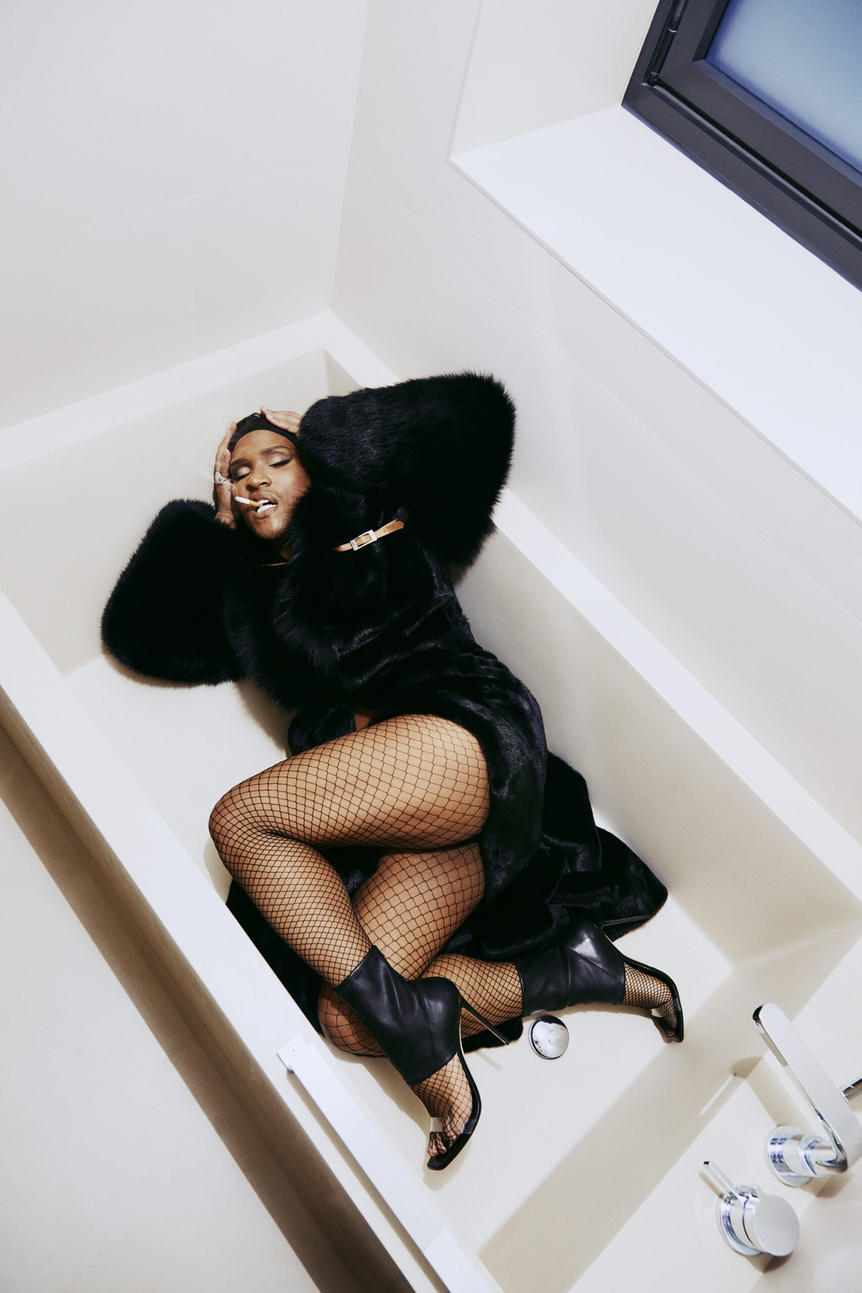 Woman in a black faux fur coat and fishnet tights lying inside a white empty bathtub