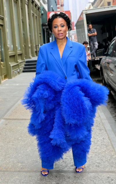 Woman in a bright blue suit and tulle pants stands on a sidewalk in an urban setting with buildings and a truck in the background.