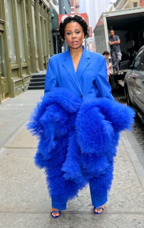 Woman in a bright blue suit and tulle pants stands on a sidewalk in an urban setting with buildings and a truck in the background.