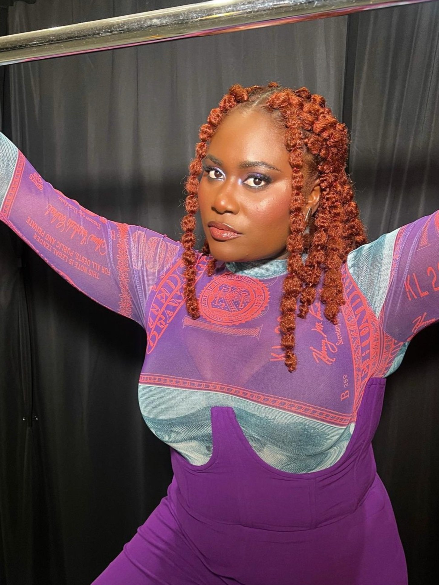 Close-up studio portrait of a Black woman with orange braided hair in a purple patterned top and pants set, looking at the camera.