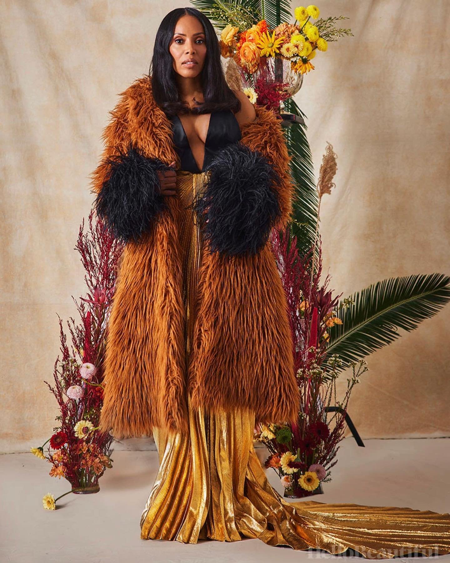 Elegant woman in fur coat poses with gold gown and floral backdrop in a studio portrait, exuding high fashion and glamour.