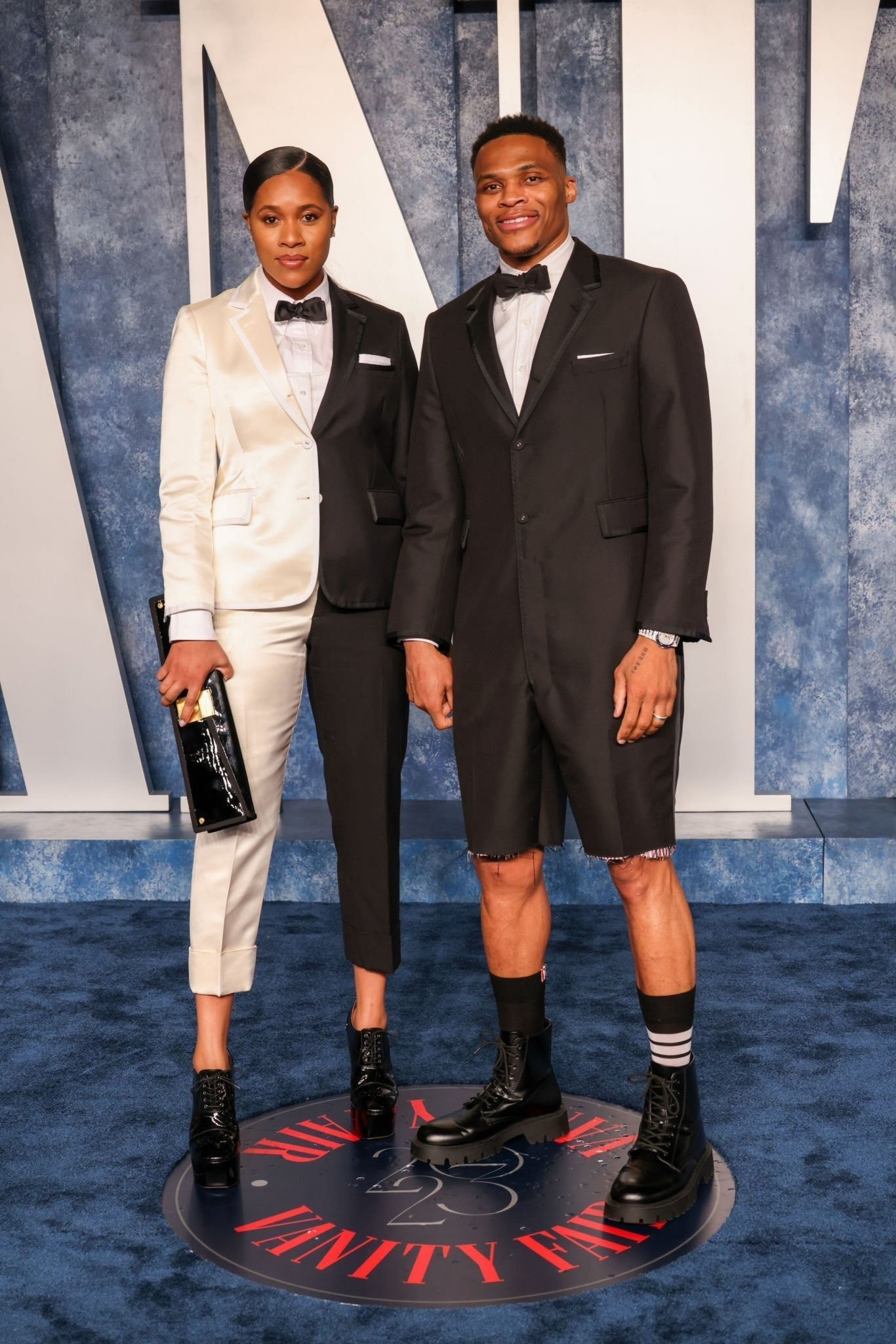 A well-dressed couple poses on a Vanity Fair red carpet; a woman in a two-tone suit and a man in a black suit and combat boots.