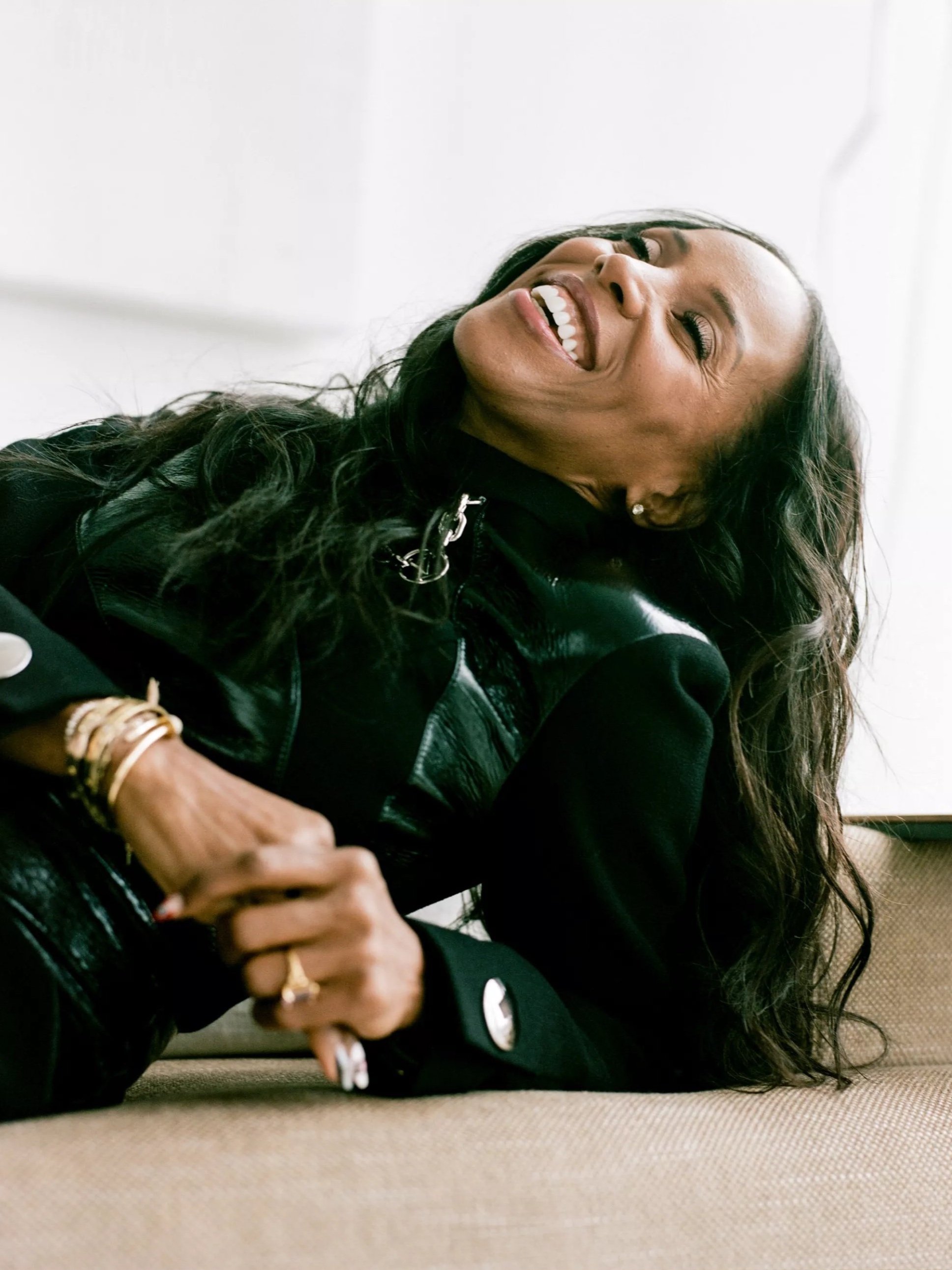 A smiling Black woman in a black jacket reclines against a neutral backdrop, wearing gold jewelry and a silver pin.