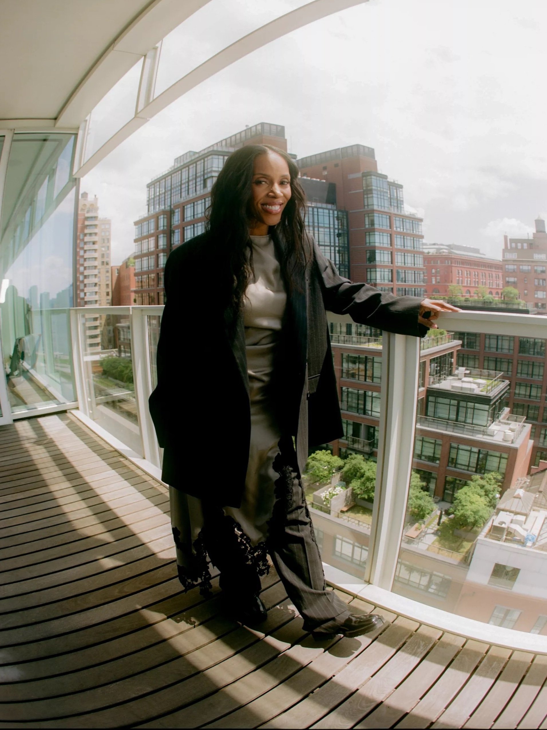 A sophisticated African-American woman in a black jacket poses on a wood-floored balcony, with city buildings visible in the background.