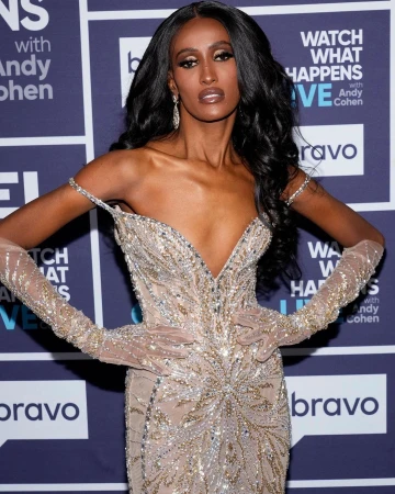 An adult woman with long dark hair poses in a champagne-colored evening gown with beaded details at an event with a purple backdrop.
