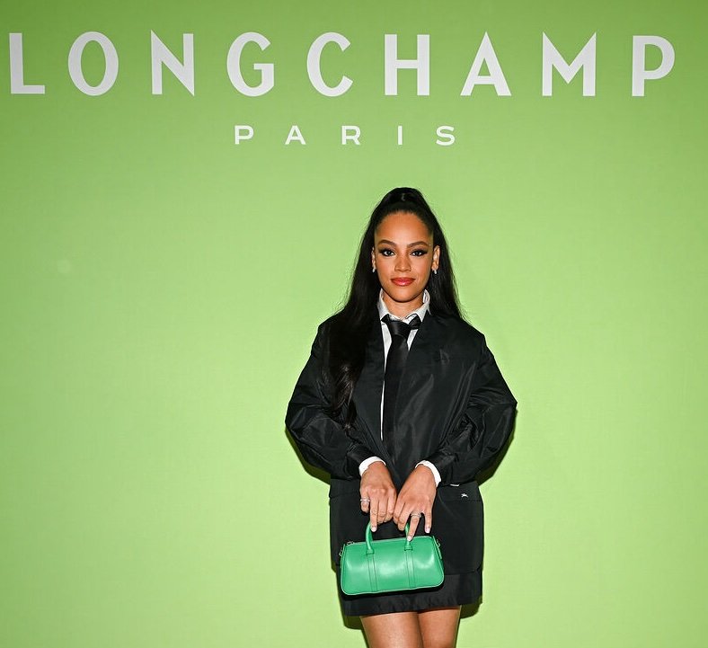 A woman in a black jacket holds a green handbag against a bright green backdrop featuring the Longchamp Paris logo.