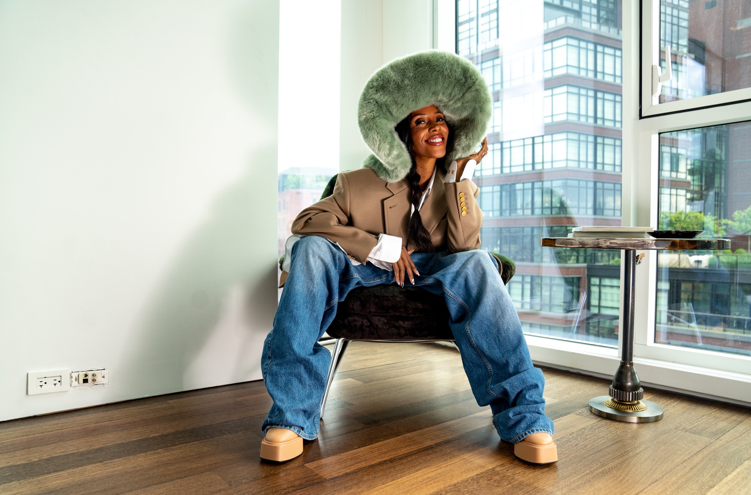 Woman in oversized jeans and statement fur hat poses indoors. Neutral blazer, platform shoes, dark hair, side table and cityscape visible.