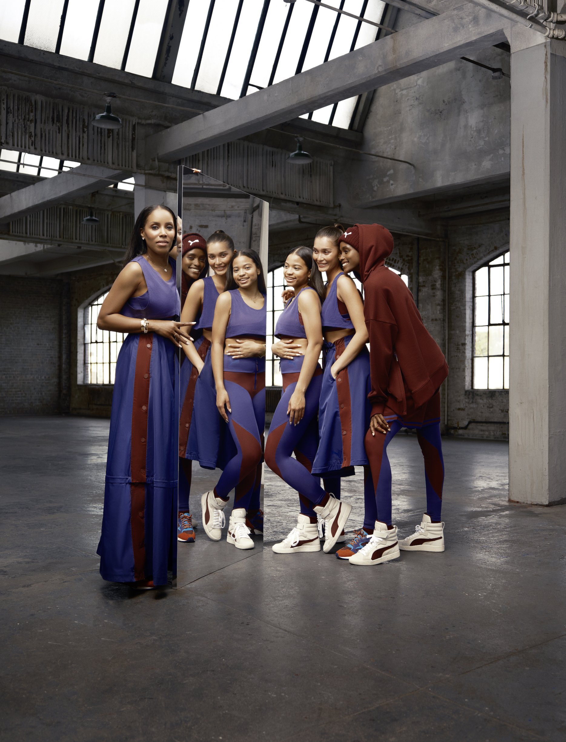 Group of women in athletic and casual clothing stand in a raw concrete space with high ceilings and natural light from large windows.