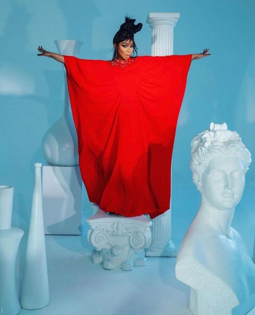 A woman in a red dress stands with outstretched arms on a white pedestal in front of a light blue backdrop with white classical busts and vases.