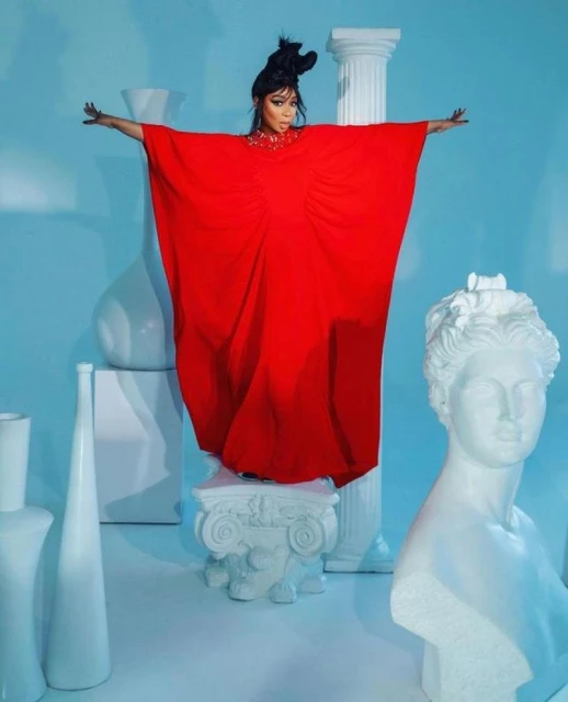 A woman in a red dress stands with outstretched arms on a white pedestal in front of a light blue backdrop with white classical busts and vases.