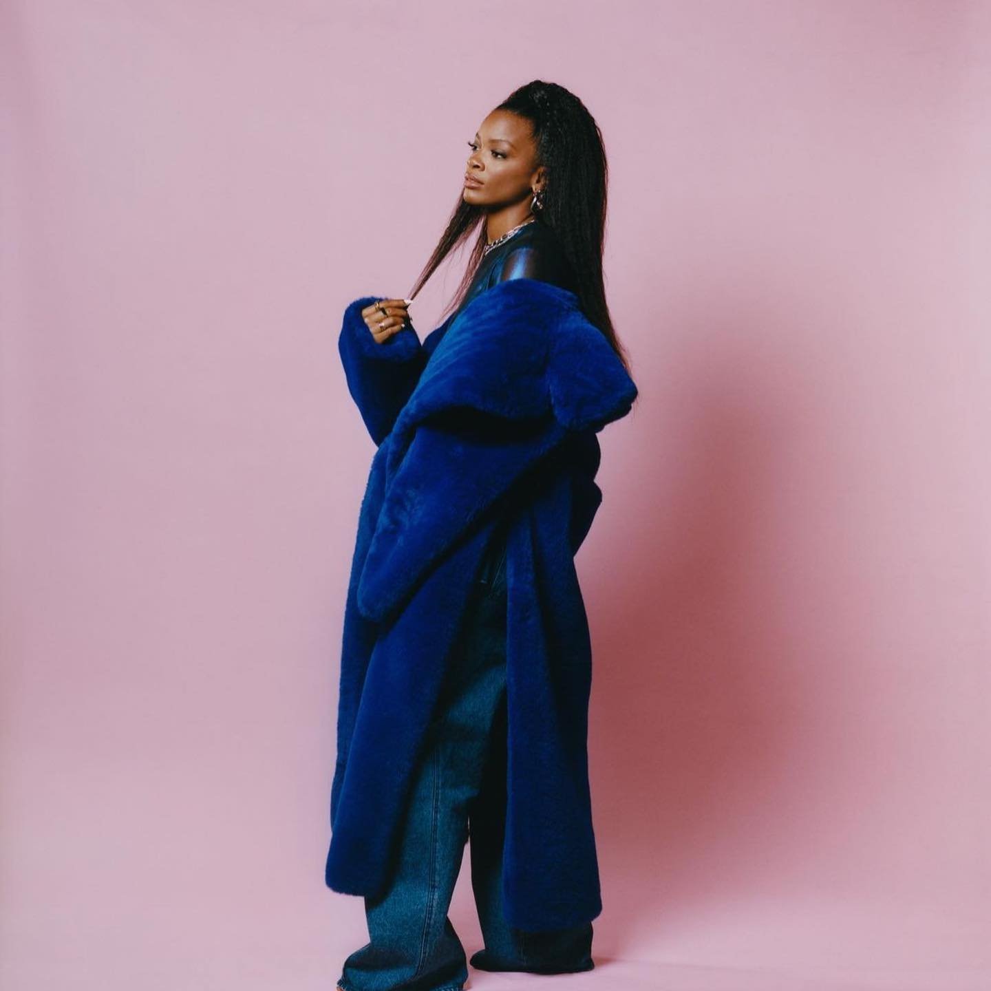 A stylish Black woman wearing a blue faux fur coat and jeans poses in a studio with a pink backdrop. Fashion editorial shot.