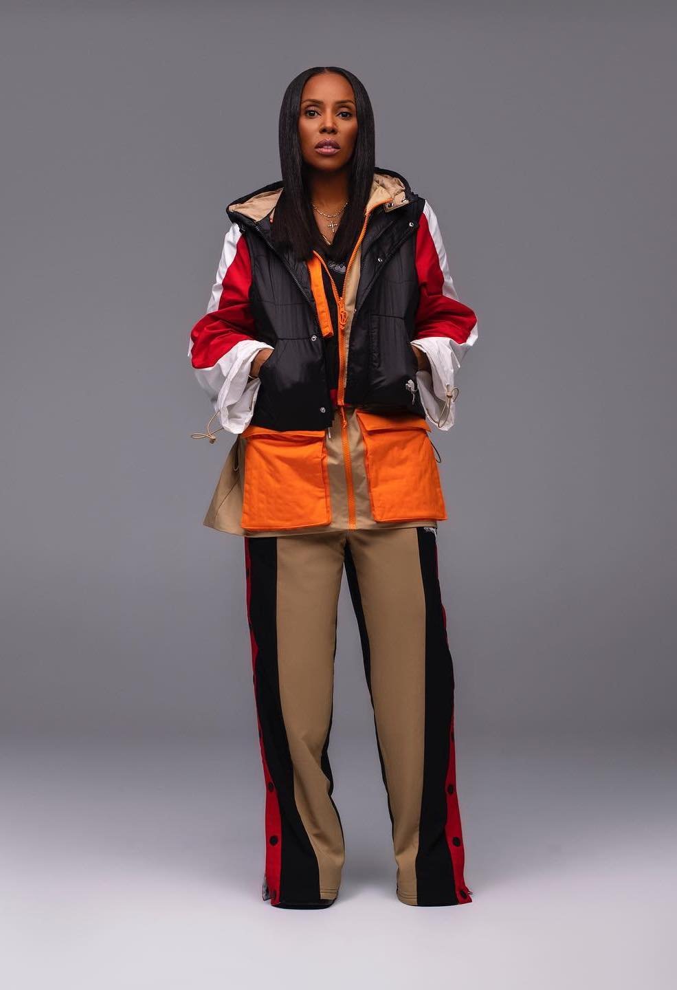Adult female in a studio shot, wearing a color-blocked vest and pants. She is holding her hands in her pockets with a neutral facial expression.