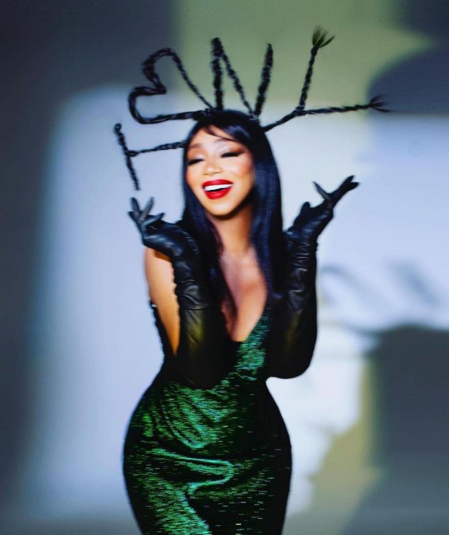 A woman poses wearing a green sequined dress and opera gloves, with braided black hair styled into antennae shapes in a studio setting.