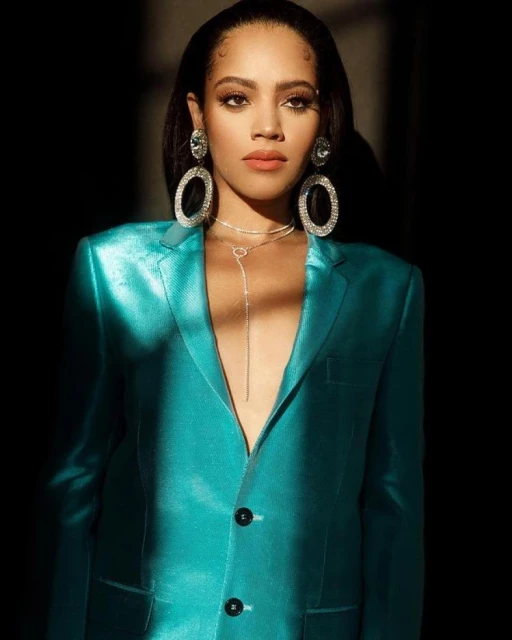 Elegant woman in a teal blazer with large, sparkling earrings and necklace posing against a dark background in a studio shot.