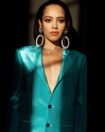 Elegant woman in a teal blazer with large, sparkling earrings and necklace posing against a dark background in a studio shot.