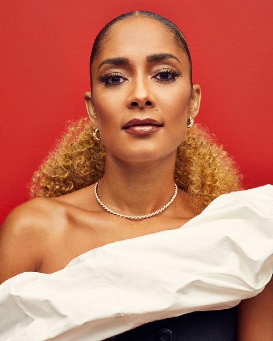 Portrait of a woman with blonde curly hair, wearing a white ruffled top, posing against a vibrant red backdrop in a photo studio.