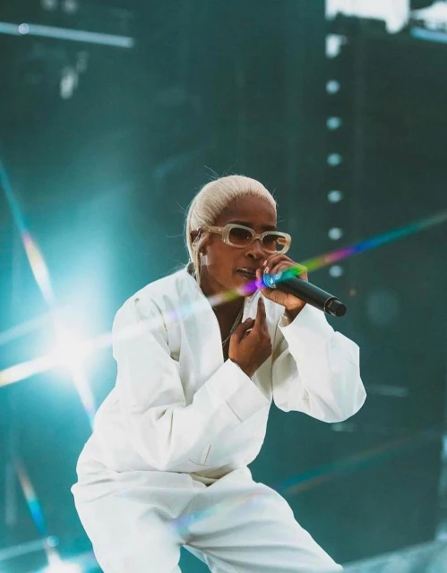 Singer performing on stage with a microphone wearing sunglasses and a white jumpsuit against a teal background with a light flare.