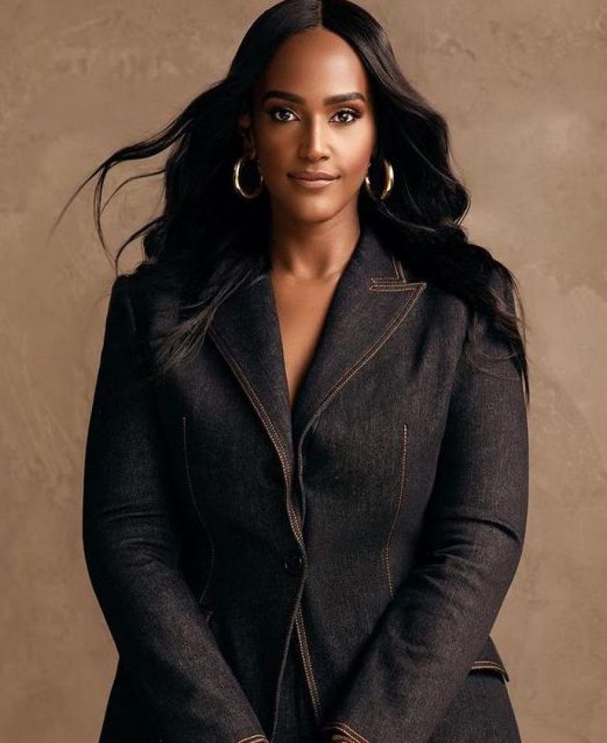 Portrait of an adult woman with long dark hair wearing a dark blue denim suit, posing in front of a neutral tan background.
